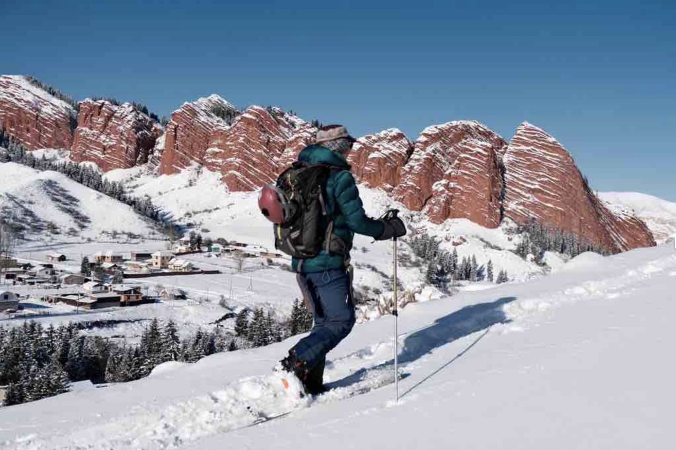 Splitboarder climbing a snowy slope below the red Seven Bulls cliffs, village in the valley
