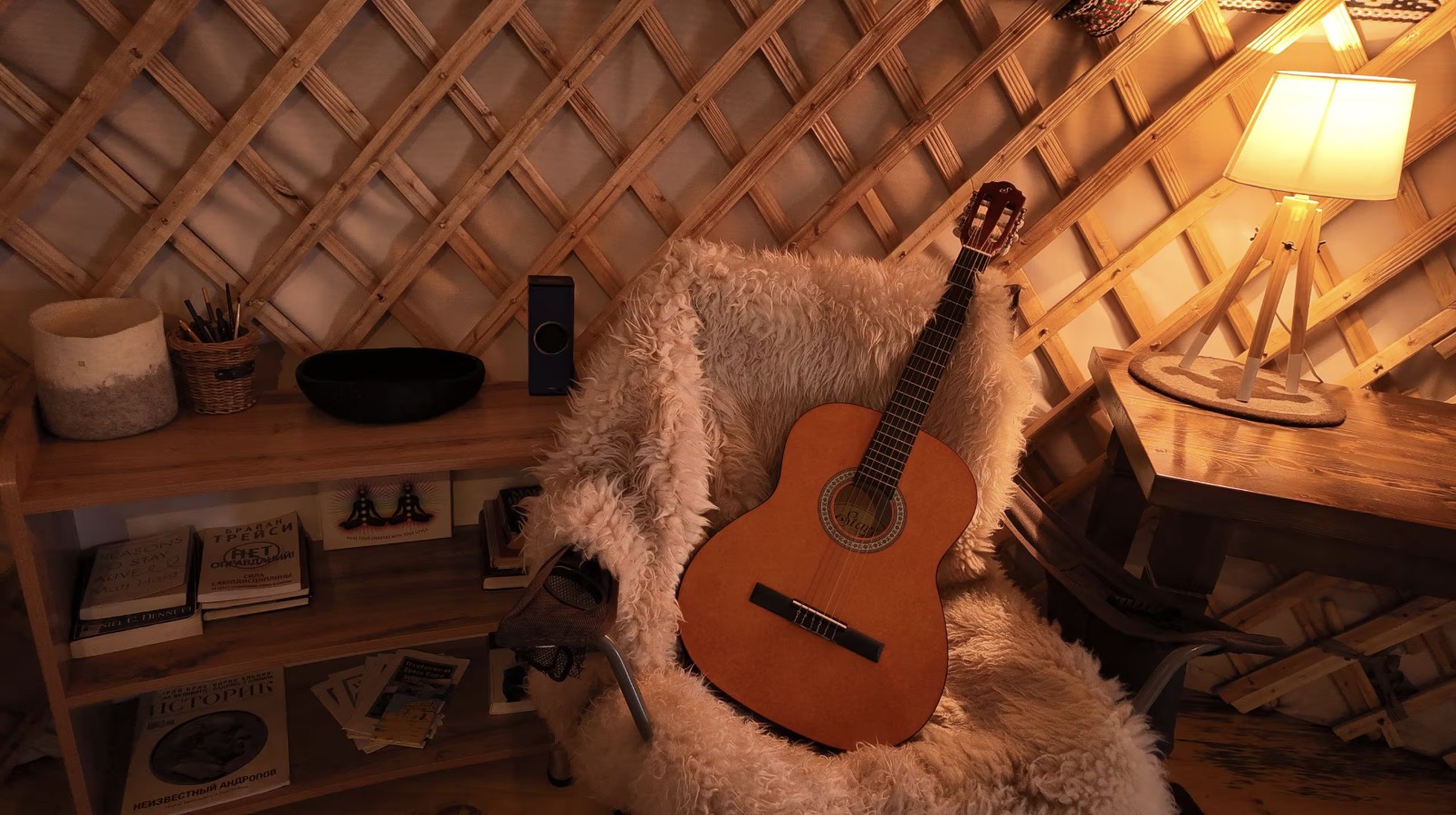 A classical guitar resting on the sheepskin chair beside a warm tripod lamp inside the yurt